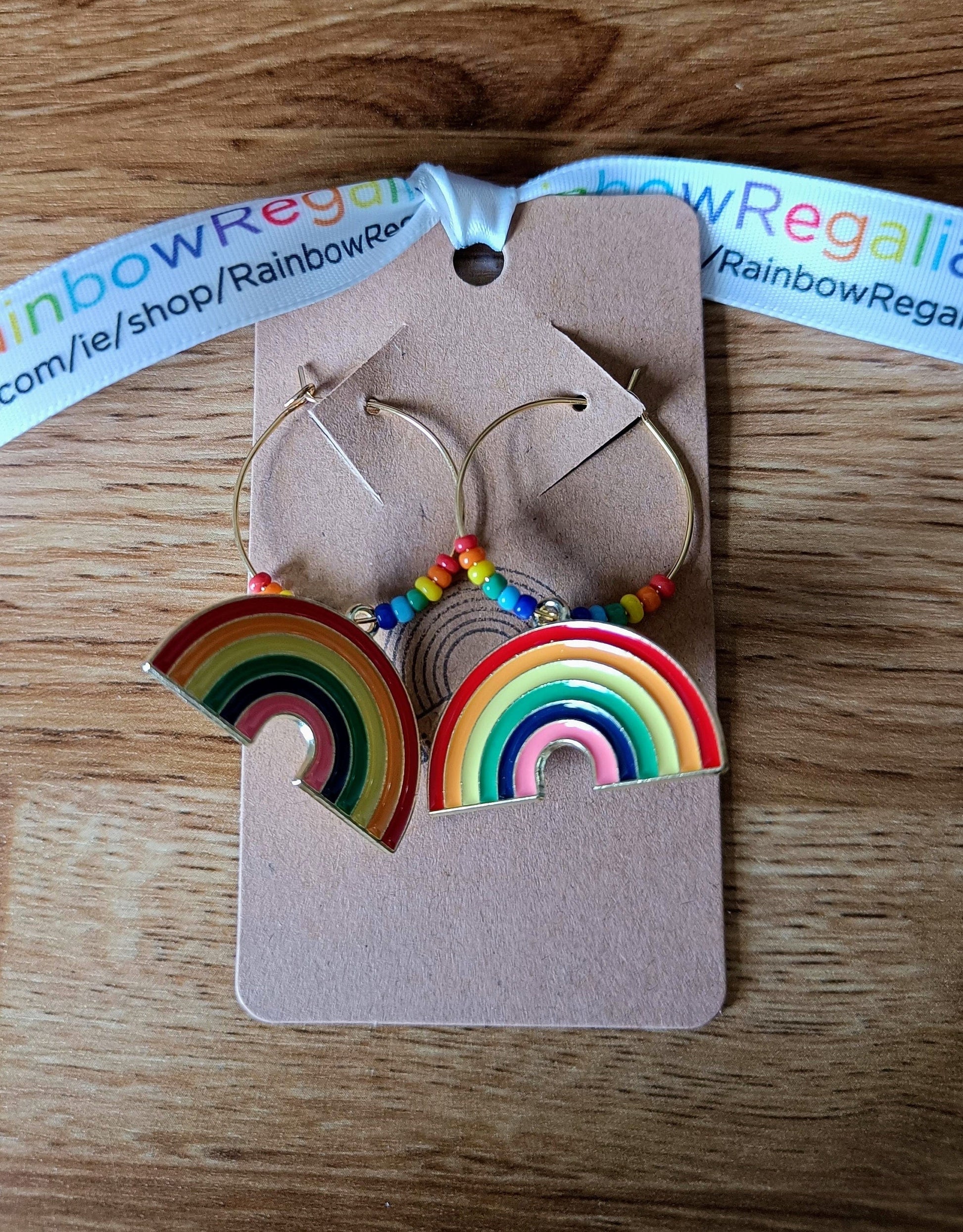 Rainbow Hoops earrings with gold-coloured brass, repurposed glass seed beads, and enamelled rainbow charms on brown display card.