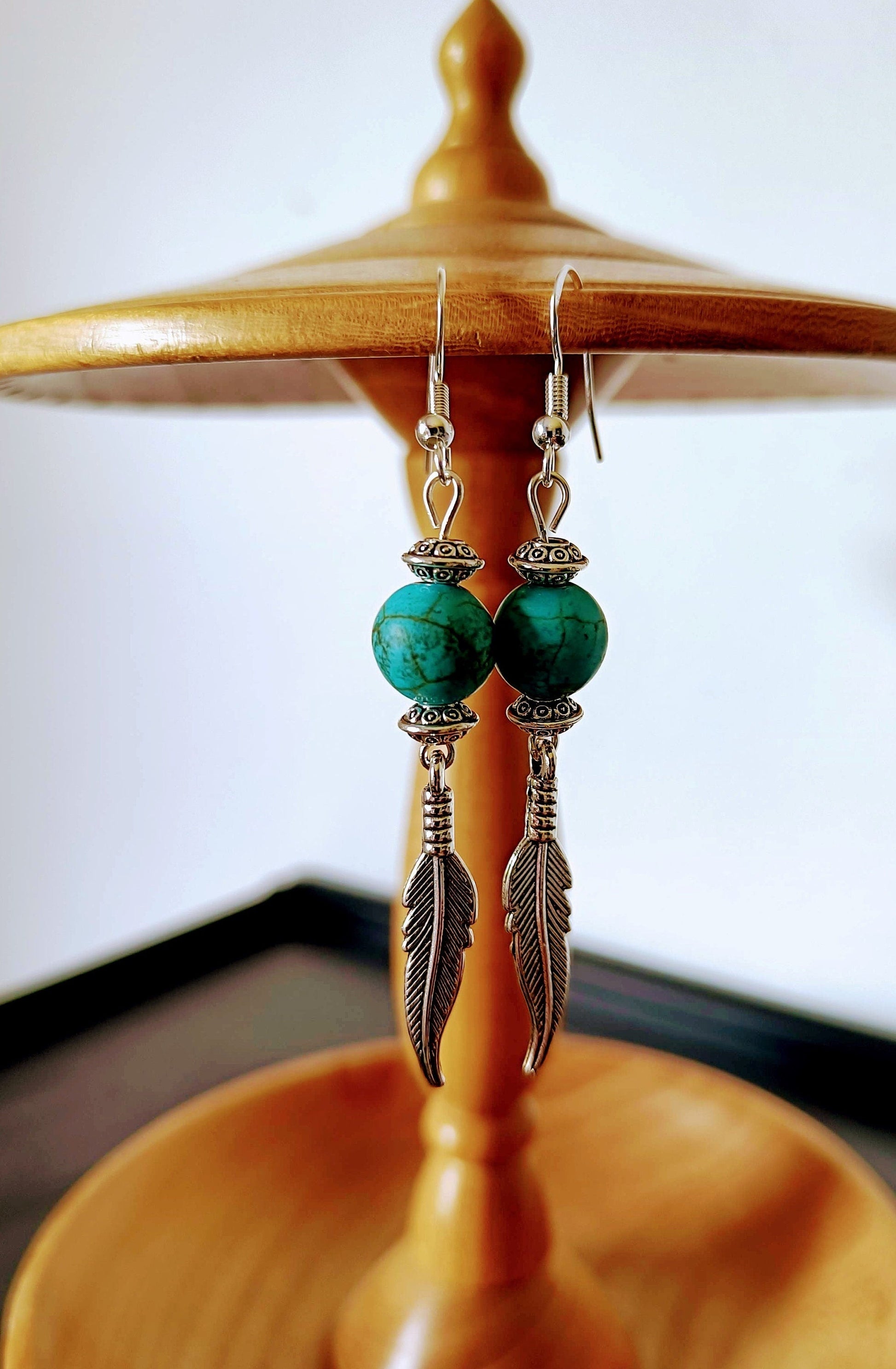 Tallulah Feather Earrings with turquoise beads and antique silver feathers hanging on a wooden stand.