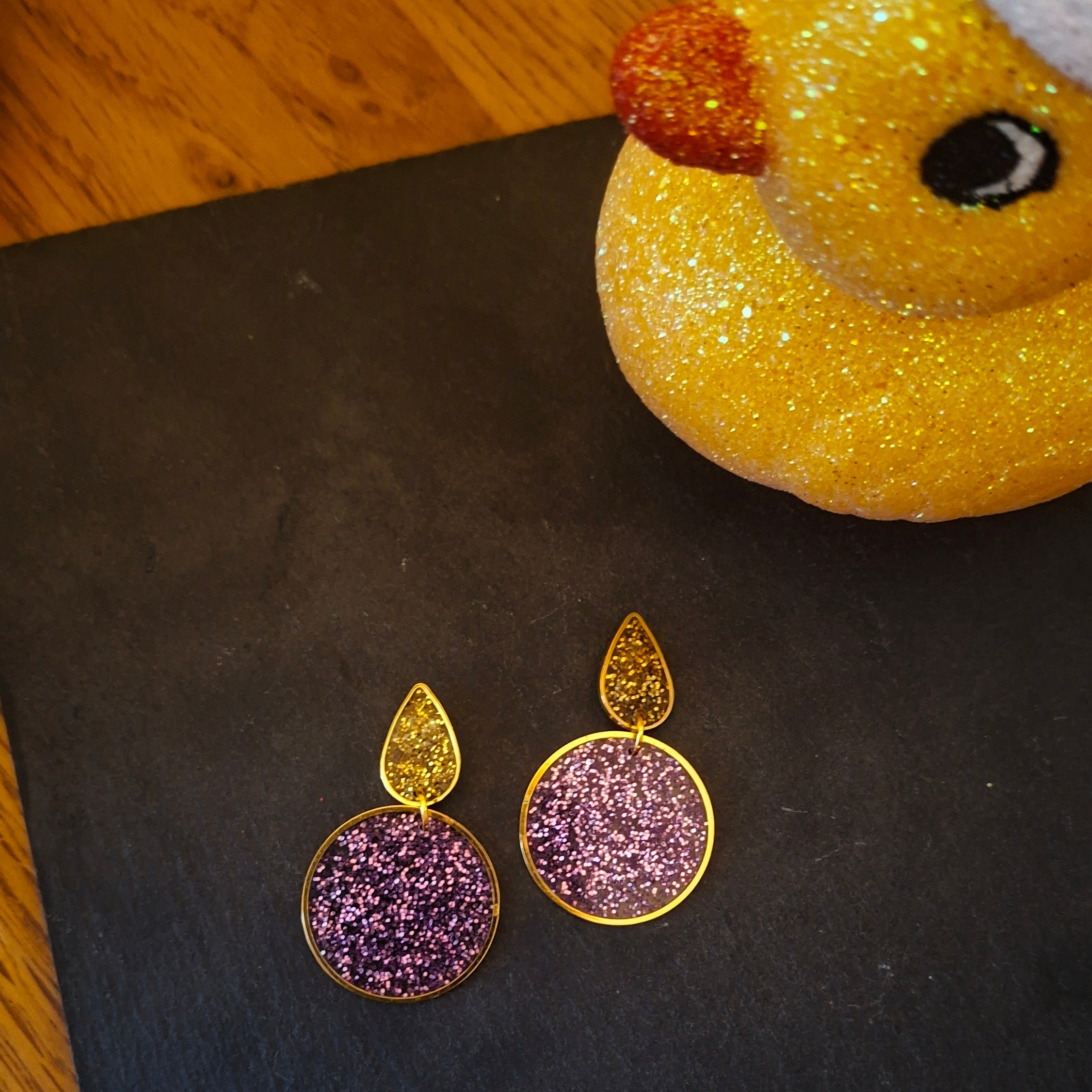 Gold and purple glittery earrings on a black surface with a yellow rubber duck in the background.
