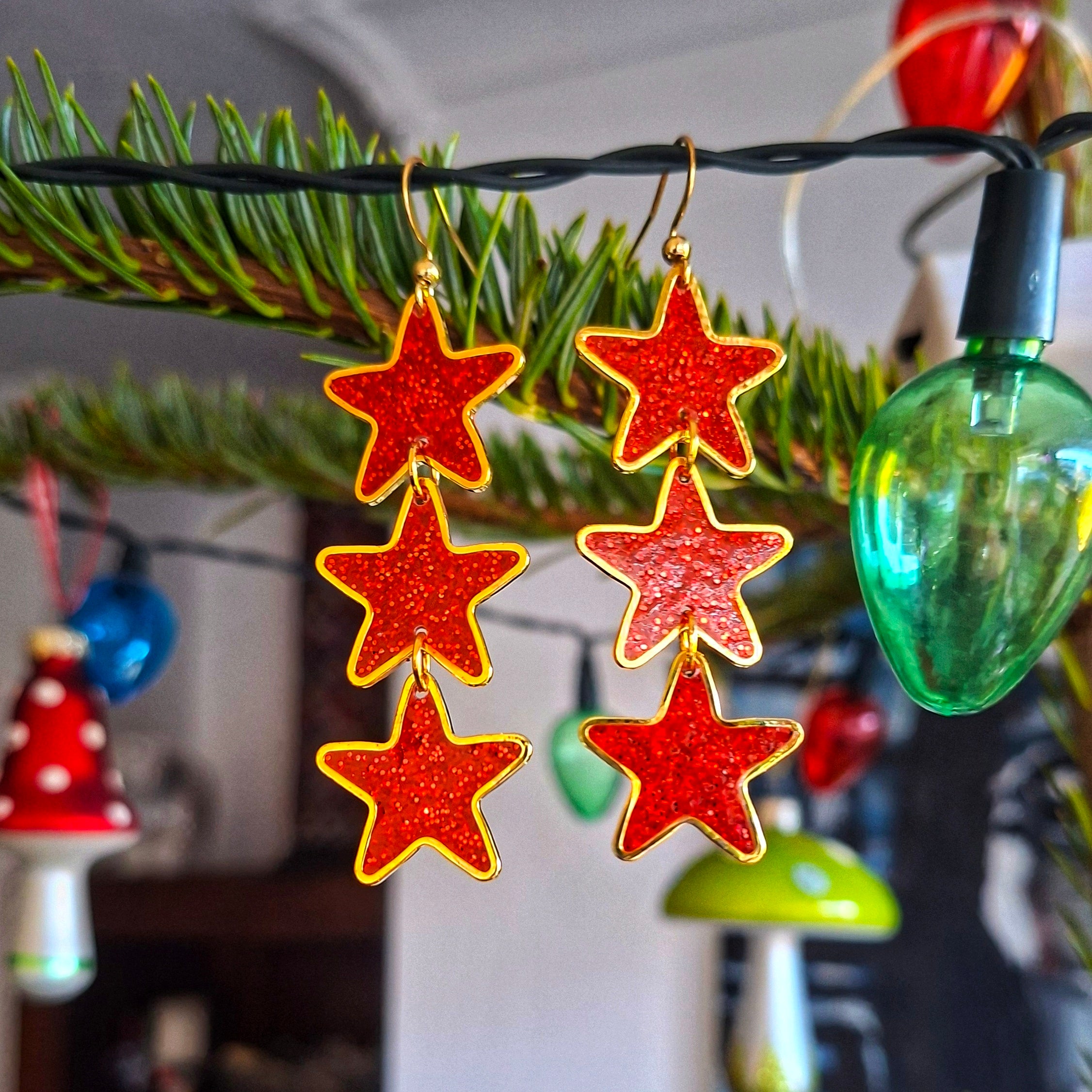 Red star-shaped earrings hanging on a string with Christmas decorations in the background.