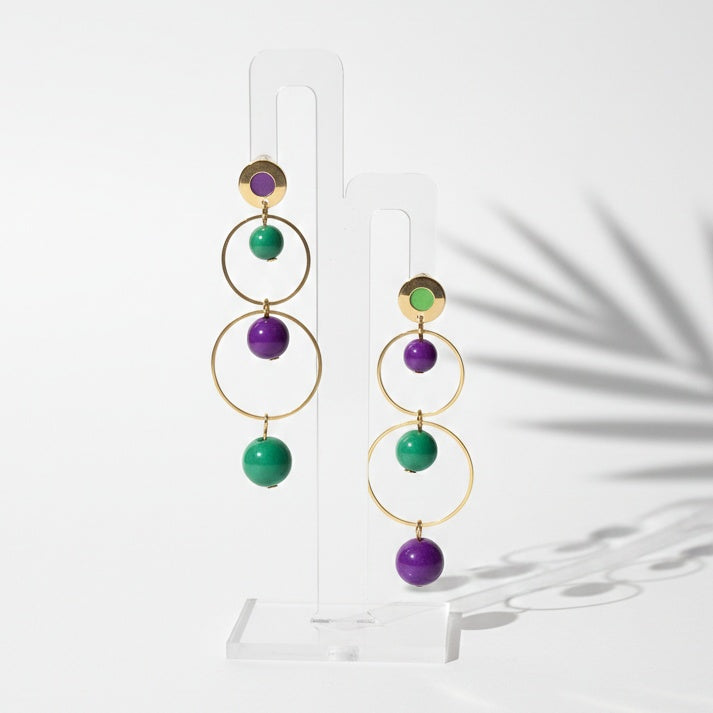 Pair of gold hoop earrings with green and purple beads on a white stand. Handmade by RainbowRegalia, Greystones, Co. Wicklow.