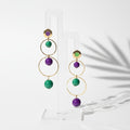 Pair of gold hoop earrings with green and purple beads on a white stand. Handmade by RainbowRegalia, Greystones, Co. Wicklow.