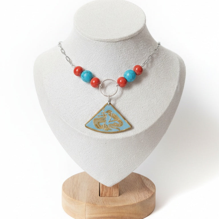A fan-shaped pendant with a gold-etched design of two ancient figures, flanked by repurposed turquoise and coral-coloured ceramic beads. 