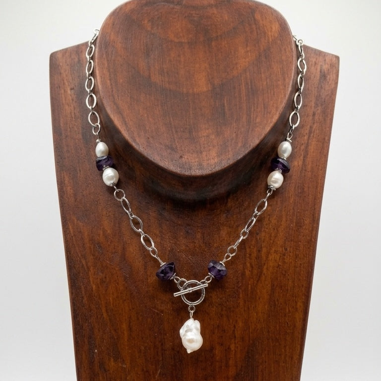 Beautiful Ruin necklace displayed on a wooden stand, featuring a baroque pearl pendant, purple amethyst beads, freshwater pearls, and a front-facing toggle clasp on a stainless steel chain.