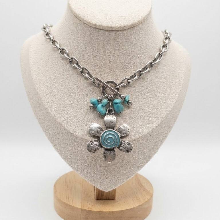 Turquoise Tempest necklace with a vintage silver flower pendant, blue spiral resin centre, turquoise stone accents, and a front toggle clasp on a stainless steel chain, displayed on a white background.