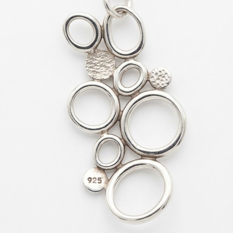A sterling silver abstract pendant featuring a cluster of interlocking open rings and textured circular charms, with a '925' silver purity stamp visible on one charm, set against a plain white background.