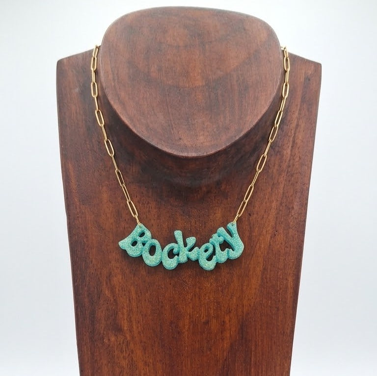 Golden green glitter ‘Bockety’ resin word pendant on a gold paperclip chain, displayed on a brown wooden necklace stand.