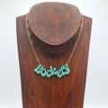 Golden green glitter ‘Bockety’ resin word pendant on a gold paperclip chain, displayed on a brown wooden necklace stand.