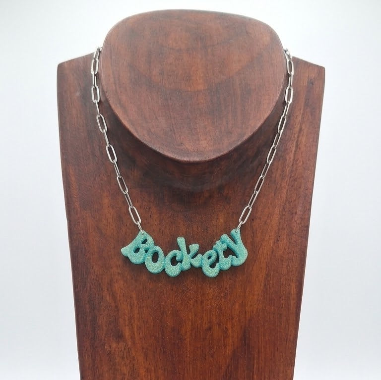 Golden green glitter ‘Bockety’ resin word pendant on a stainless steel paperclip chain, displayed on a brown wooden necklace stand.