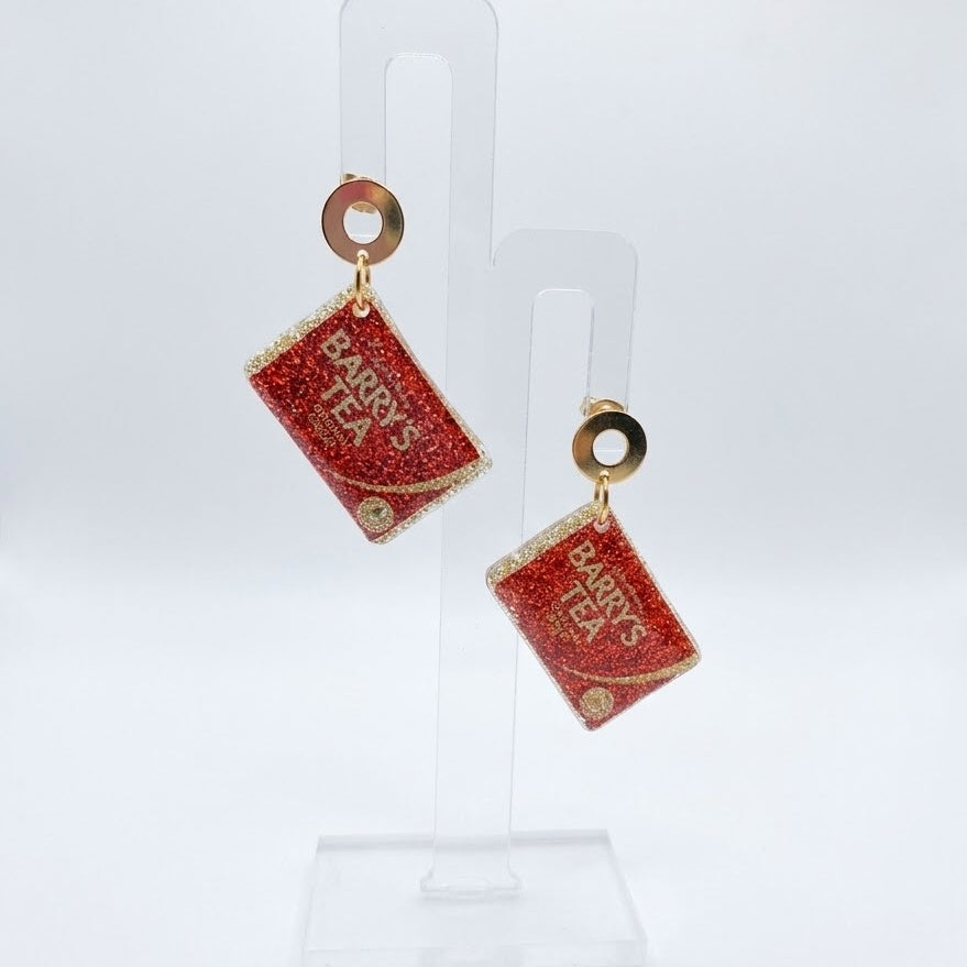 The National Brew earrings shaped like red glitter Barry’s Tea boxes, hanging from halo studs on a clear display stand against a white background.