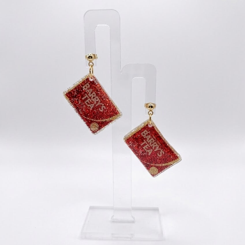 The National Brew earrings shaped like red glitter Barry’s Tea boxes, hanging from ball studs on a clear display stand against a white background.