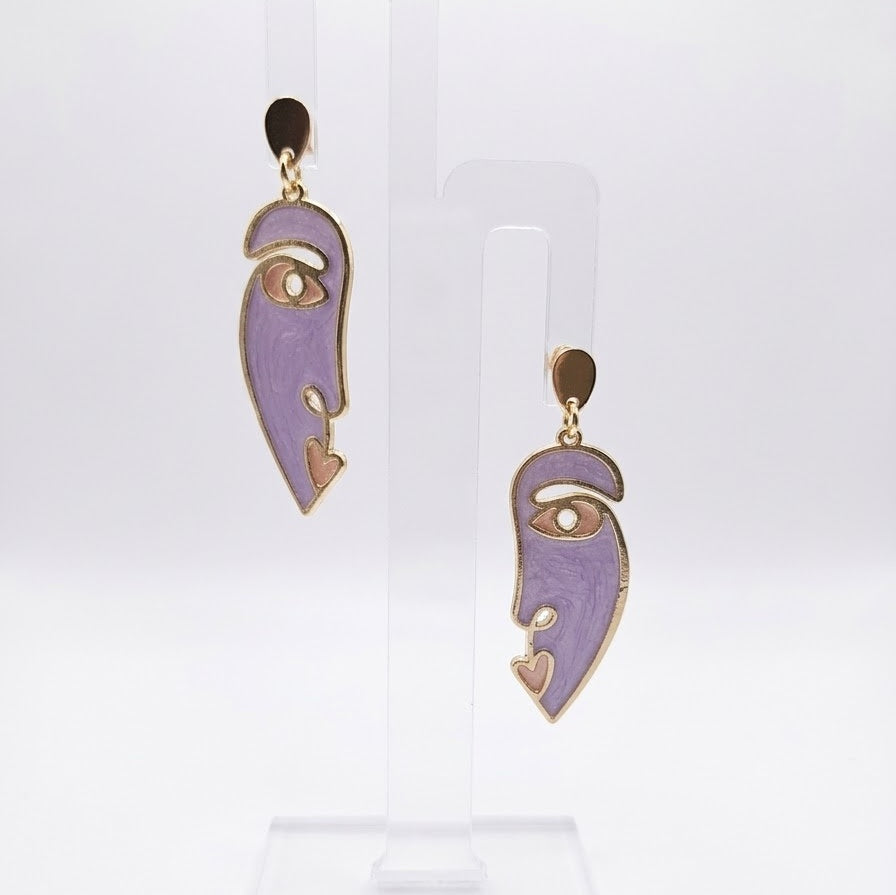 Side Eye abstract face earrings in lilac purple resin with gold-tone outlines and teardrop studs, displayed on a clear stand against white.