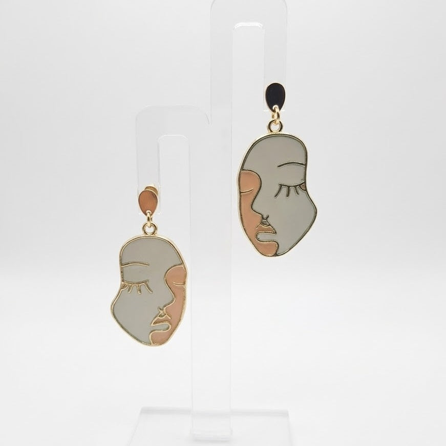 Side Eye abstract face earrings in sage green resin with gold-tone outlines and teardrop studs, displayed on a clear stand against white.