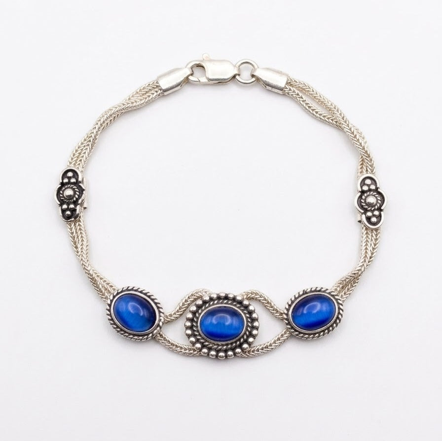 Sterling silver woven chain bracelet with three blue cat’s eye cabochons in decorative rope-edged settings and oxidised filigree accents, on a white background.