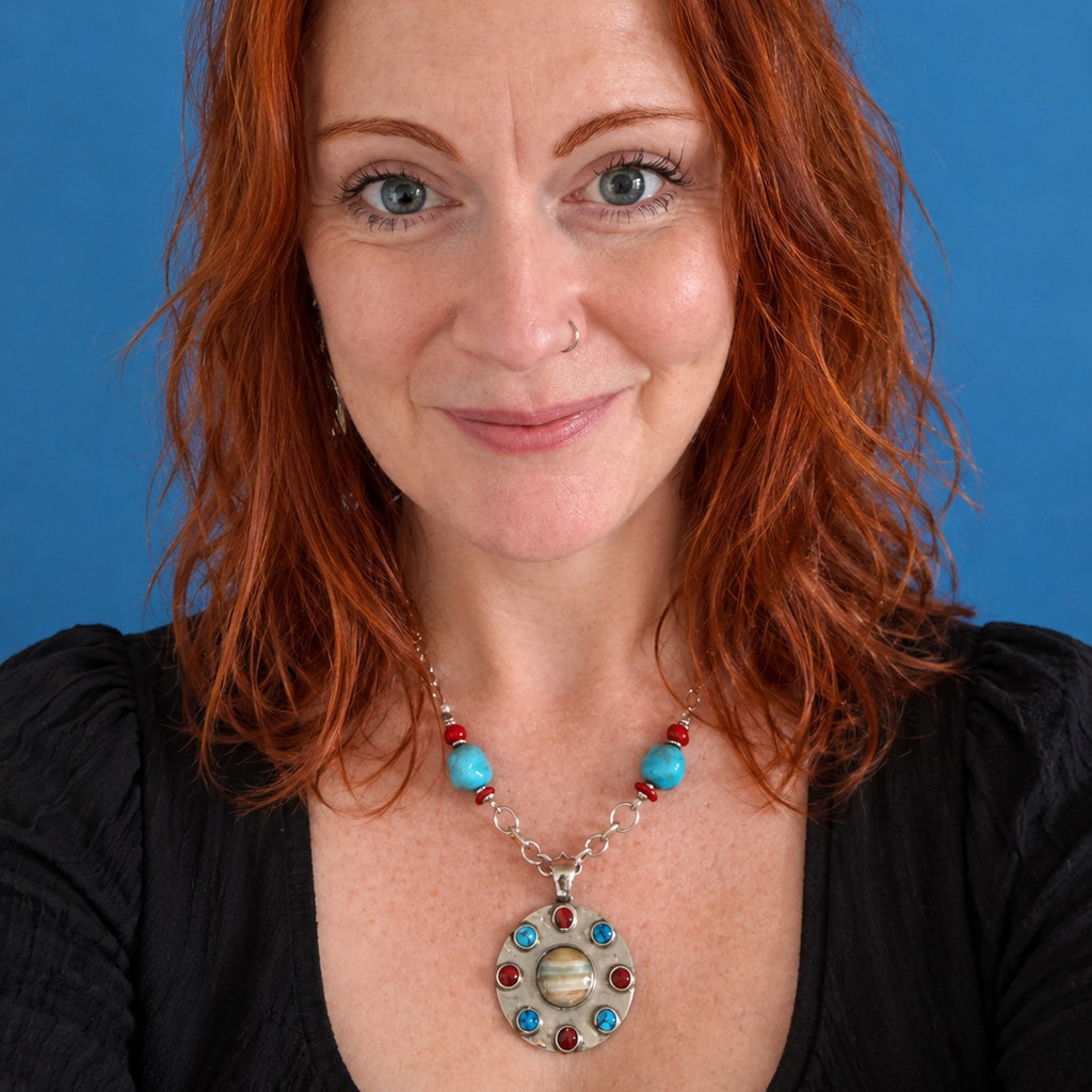 A close-up portrait of a woman with red hair wearing a silver-toned necklace with turquoise and red beads and a circular pendant featuring a planet design, set against a solid blue background.