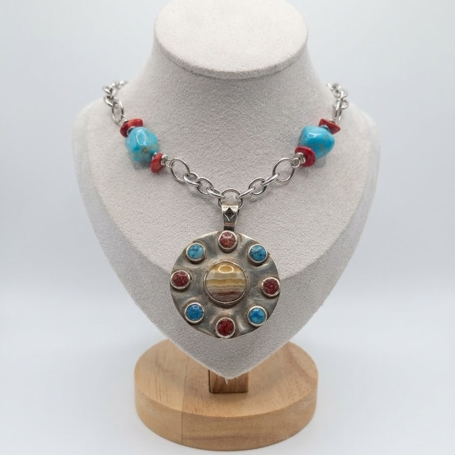 A silver chain necklace with turquoise and red beads and a circular pendant featuring sun-like stone details, displayed on a white mannequin bust with a wooden stand against a white background.
