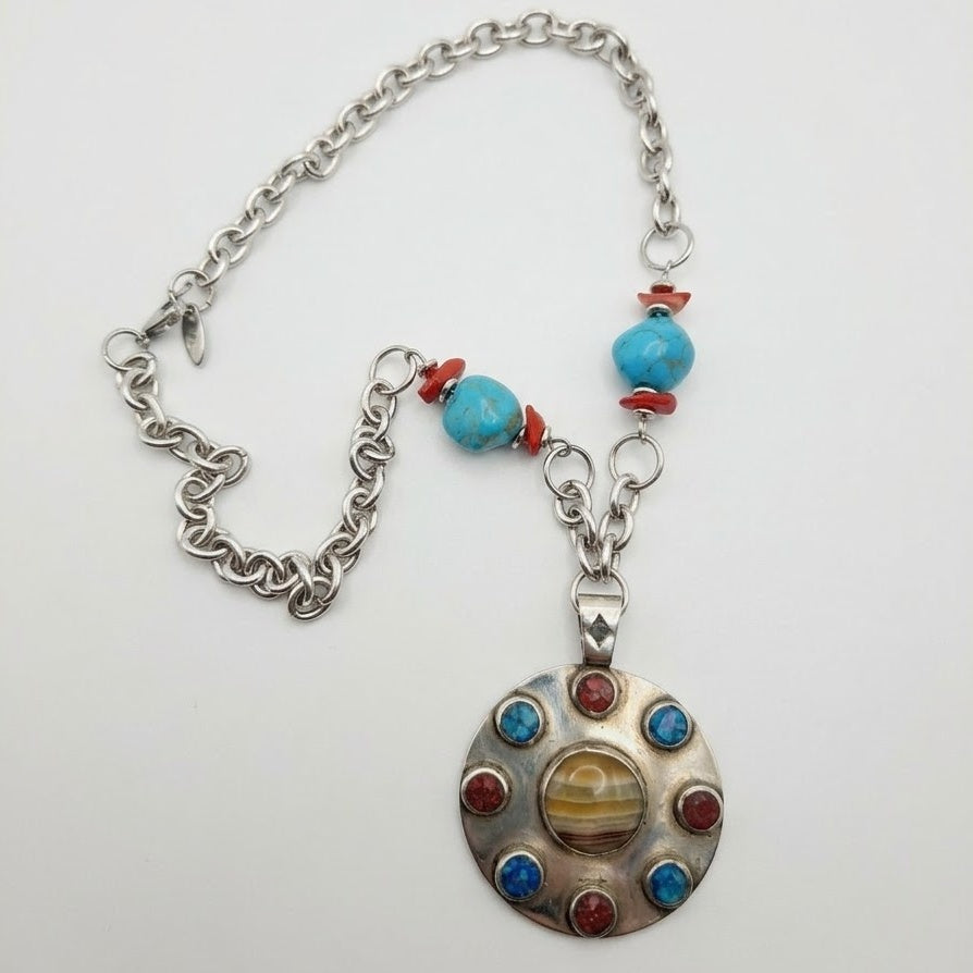 A silver-toned chain necklace with turquoise blue beads, red spacer beads, and a round silver pendant featuring a central striped amber-like inlay surrounded by small red and blue stones, shown on a plain white background.