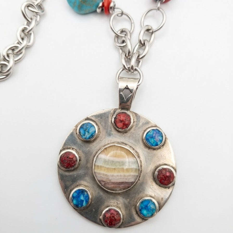 A silver-toned circular pendant necklace featuring a central oval stone with beige, brown, and cream banding, surrounded by eight bezel-set round stones in alternating red and blue, hanging from a silver chain on a white background.