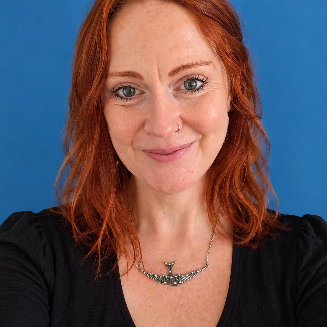 A close-up portrait of a smiling woman with red hair and freckles, wearing a silver swallow pendant necklace and a black top, set against a solid blue background.