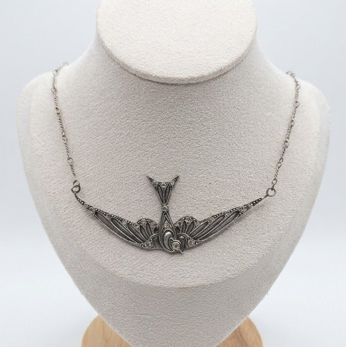 An ornate silver necklace with a swallow motif pendant, displayed on a textured white jewelry bust against a light gray background.
