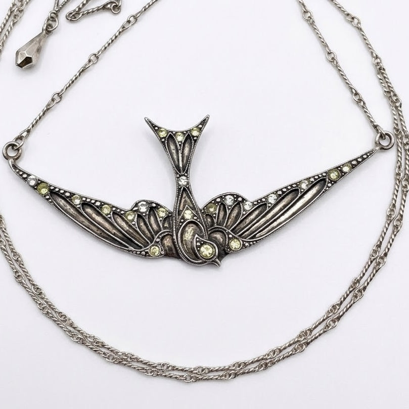 An ornate silver-toned swallow pendant necklace with intricate filigree detailing and small pale yellow gemstone accents, displayed on a clean white background.