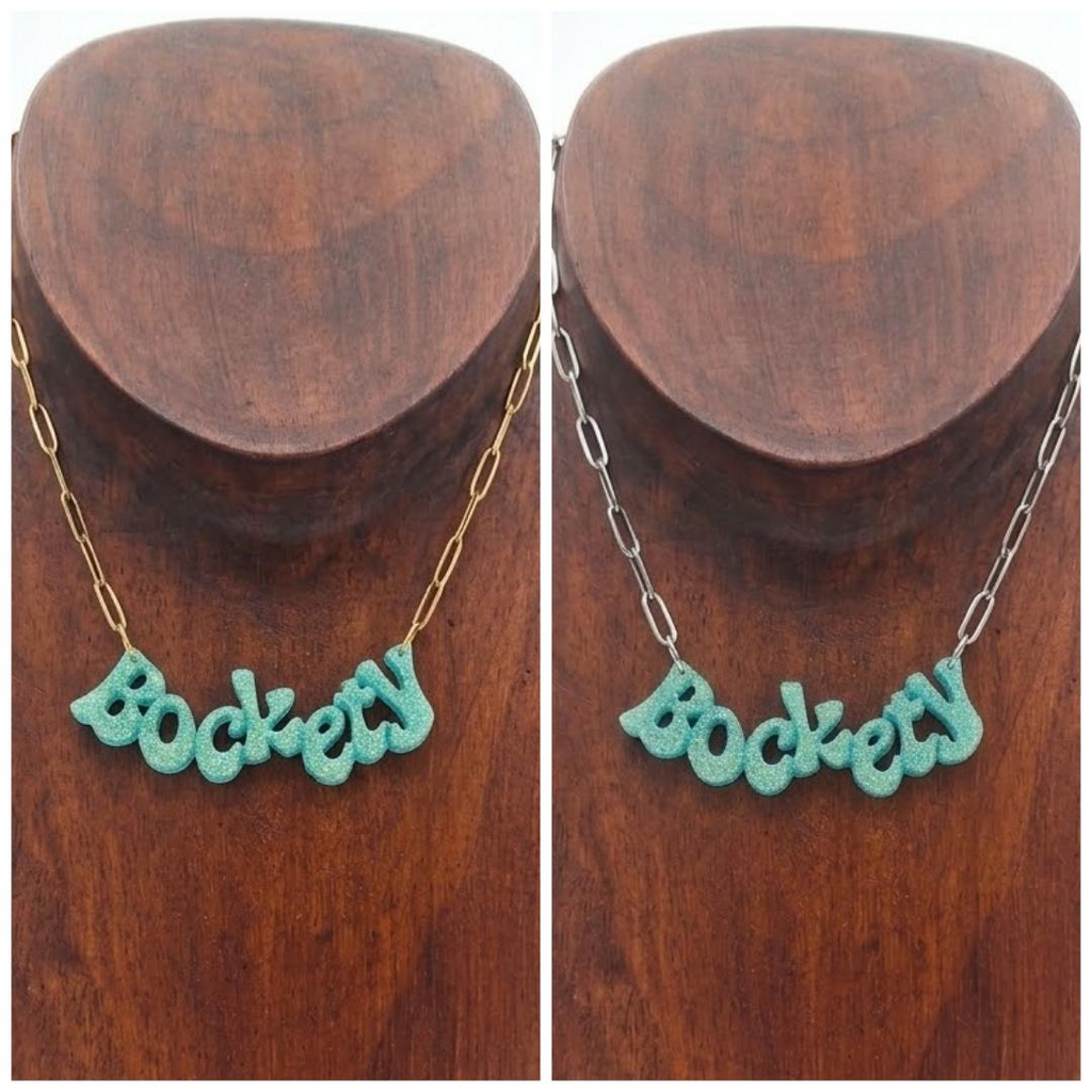 Golden green glitter ‘Bockety’ resin word pendant on a gold paperclip chain, displayed on a brown wooden necklace stand.