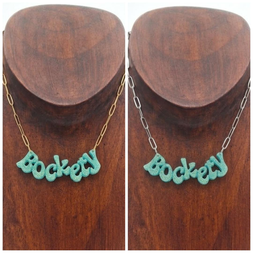 Golden green glitter ‘Bockety’ resin word pendant on a gold paperclip chain, displayed on a brown wooden necklace stand.