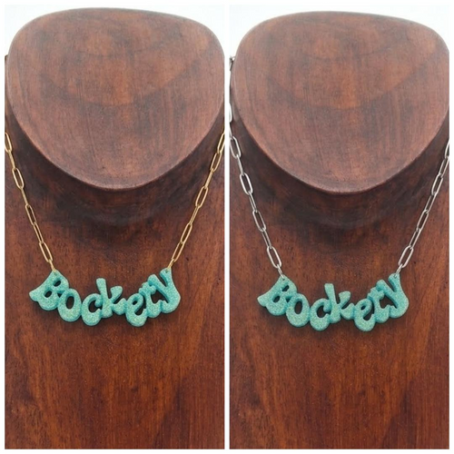 Golden green glitter ‘Bockety’ resin word pendant on a gold paperclip chain, displayed on a brown wooden necklace stand.