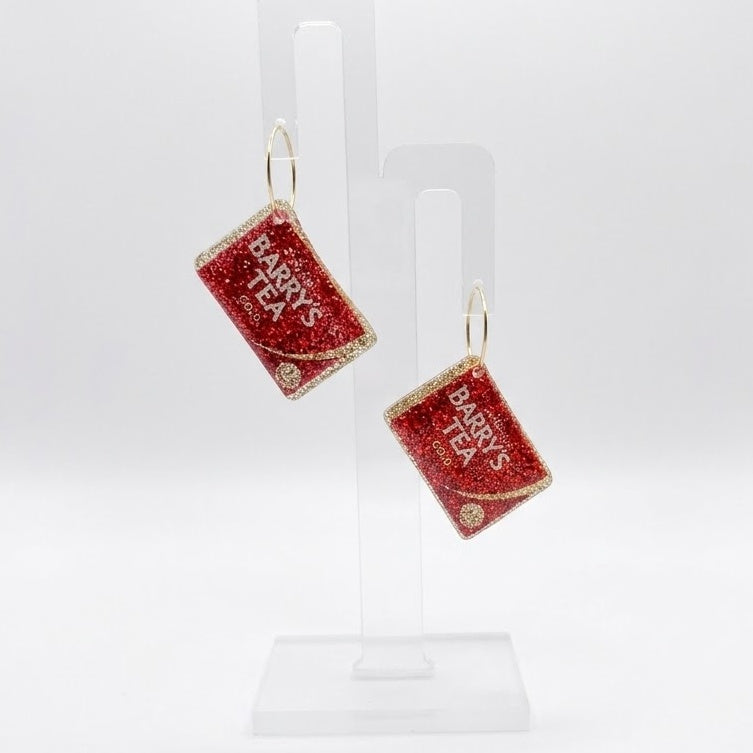The National Brew earrings shaped like red glitter Barry’s Tea boxes, hanging from hoops on a clear display stand against a white background.