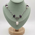Beautiful Ruin necklace displayed on a pale green bust, featuring a baroque pearl pendant, purple amethyst beads, freshwater pearls, and a front-facing toggle clasp on a stainless steel chain.