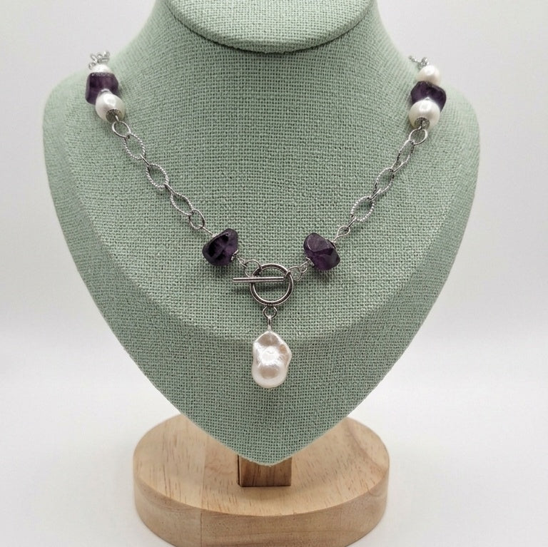 Beautiful Ruin necklace displayed on a pale green bust, featuring a baroque pearl pendant, purple amethyst beads, freshwater pearls, and a front-facing toggle clasp on a stainless steel chain.