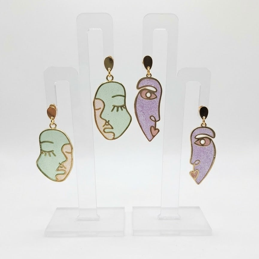Side Eye abstract face earrings in sage green and lilac purple resin with gold-tone outlines and teardrop studs, displayed on a clear stand against white.