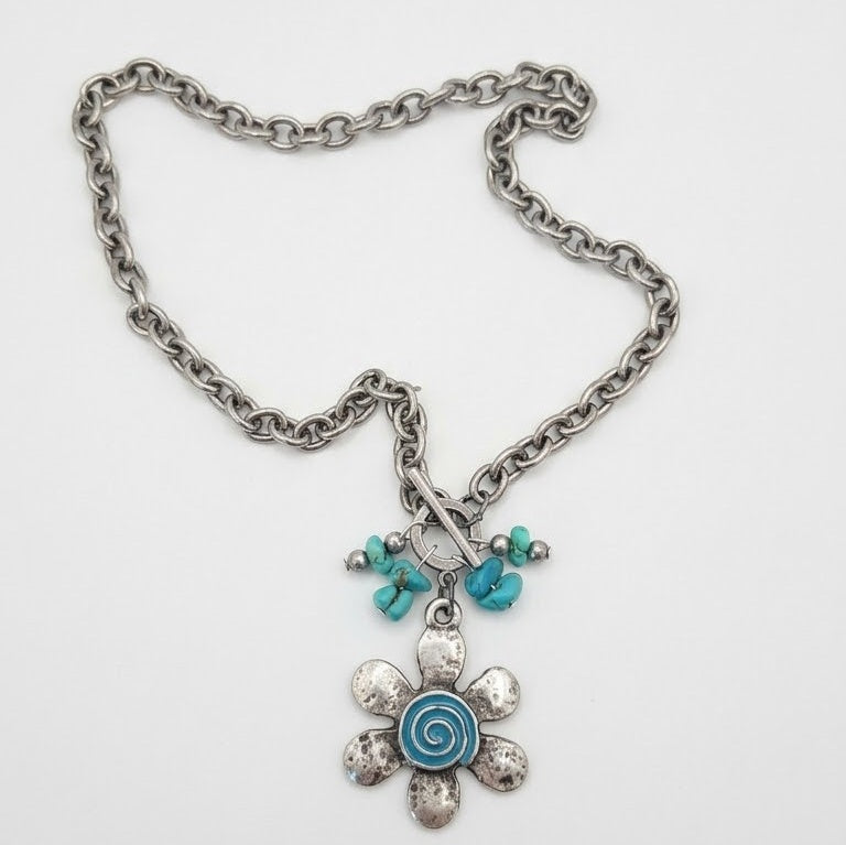 Turquoise Tempest necklace with a vintage silver flower pendant, blue spiral resin centre, turquoise stone accents, and a front toggle clasp on a stainless steel chain, displayed on a white background.
